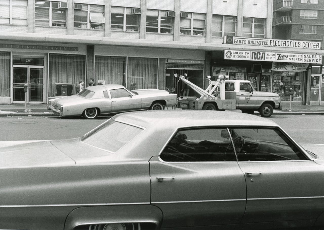 expertise-01 Black and white photo of vintage cars with a tow truck in an urban street setting.